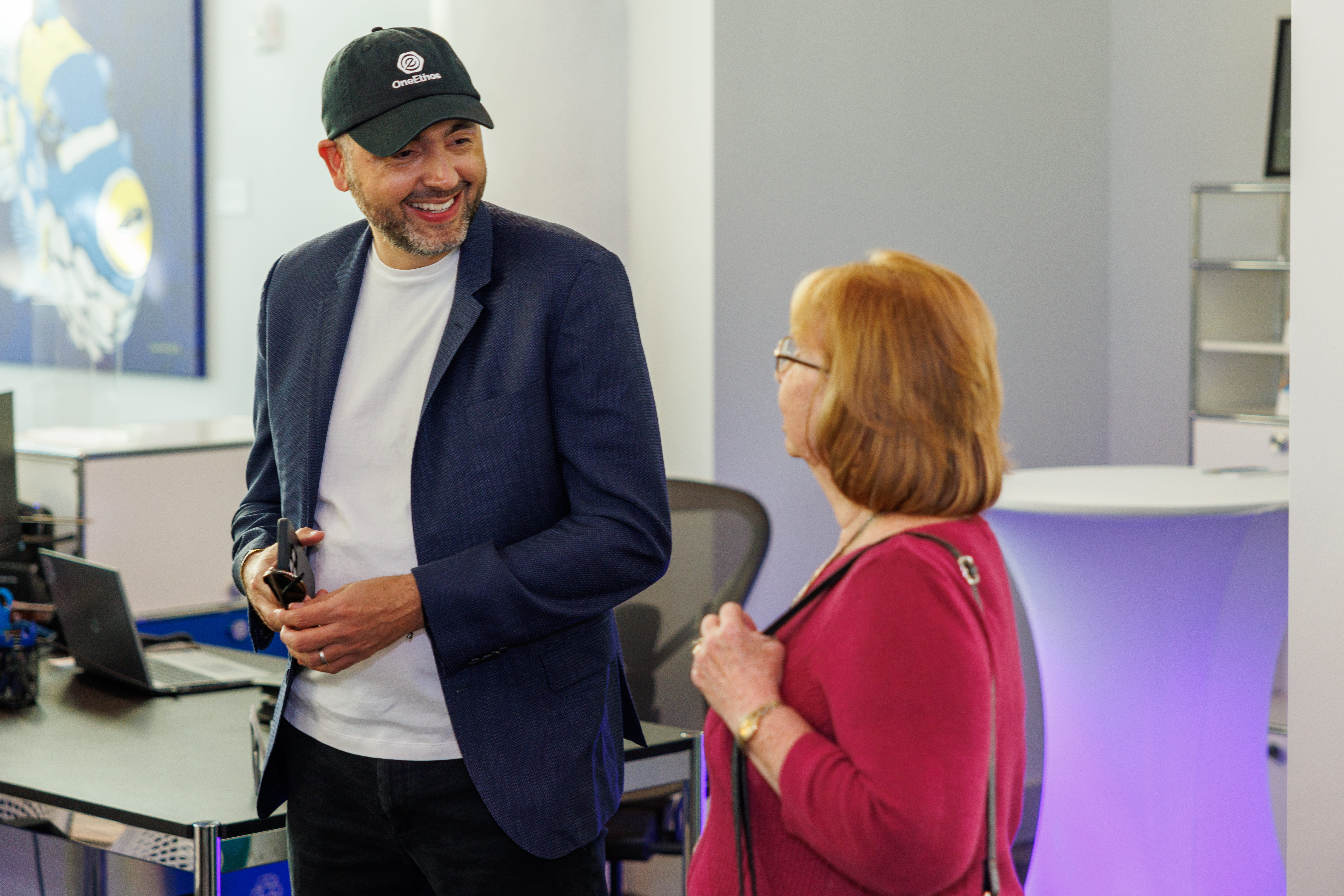 Man in a black cap and navy blazer smiling and talking to a woman in a red sweater in an office setting.
