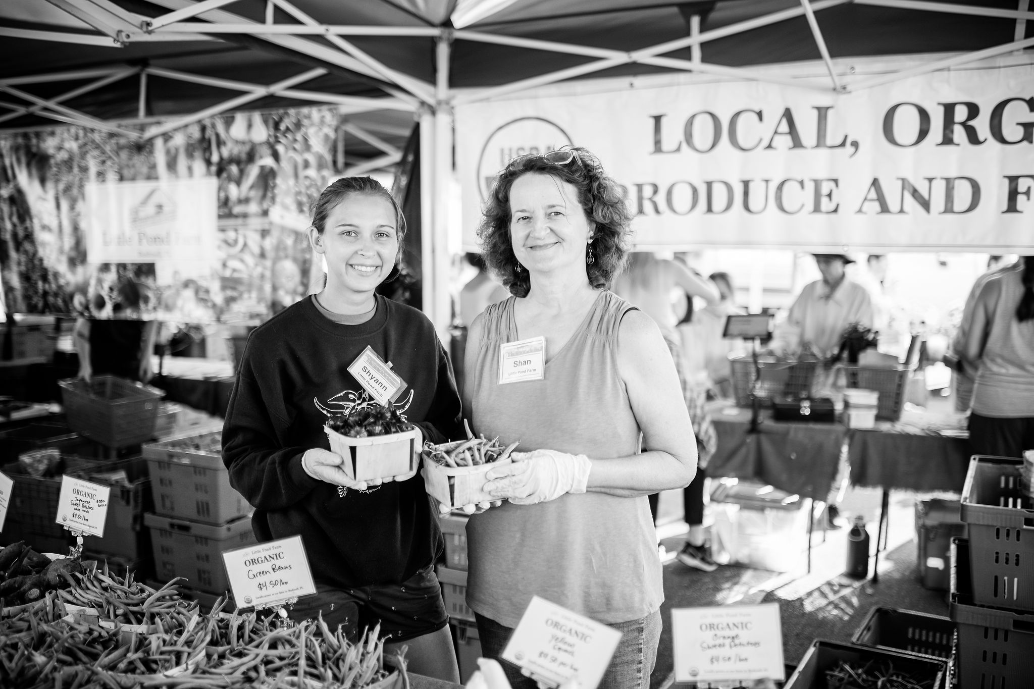 Two women at market stall holding baskets of produce, with organic produce signs visible.