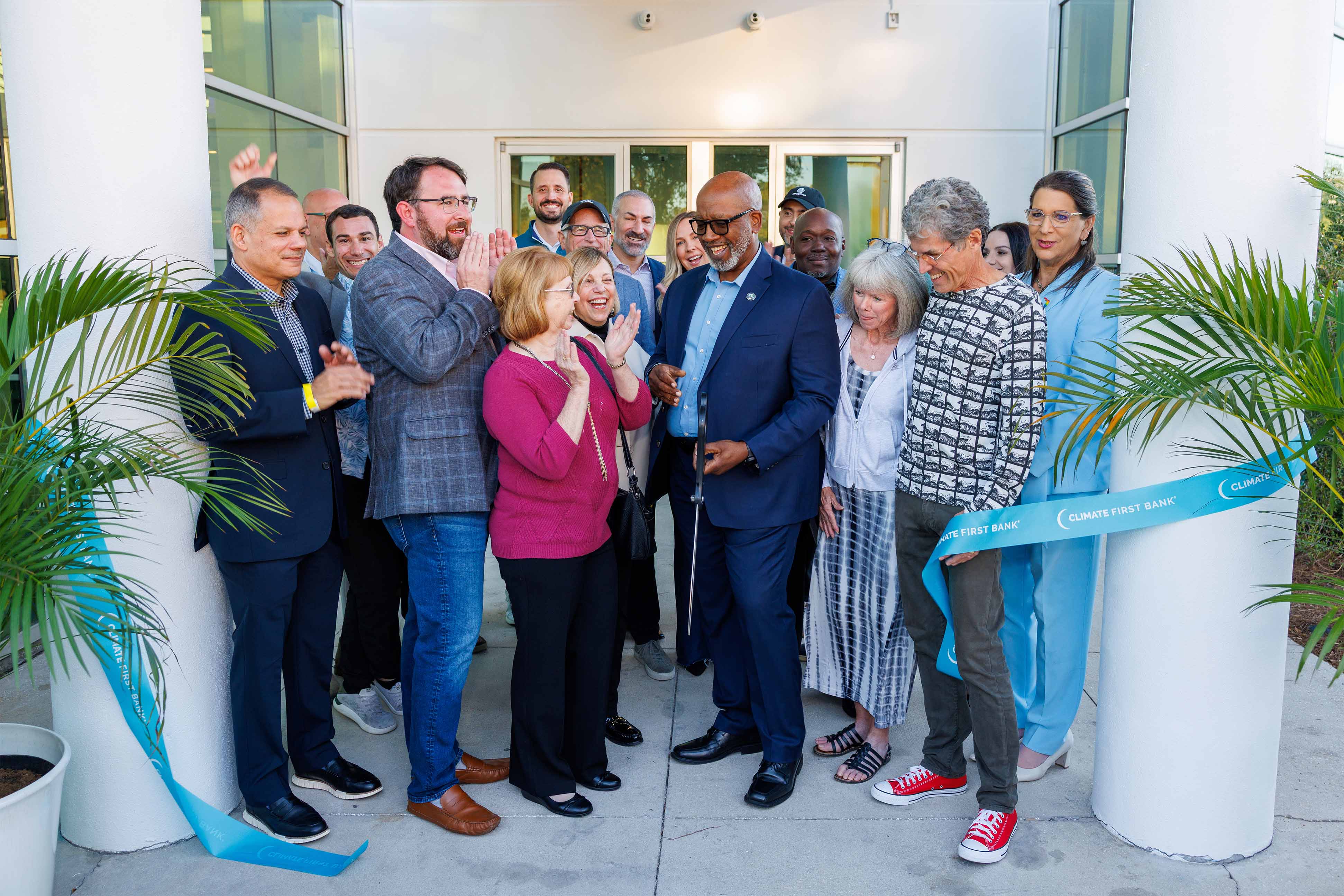Group of people gathered outside a building for a ribbon-cutting ceremony with a man in a suit holding scissors.