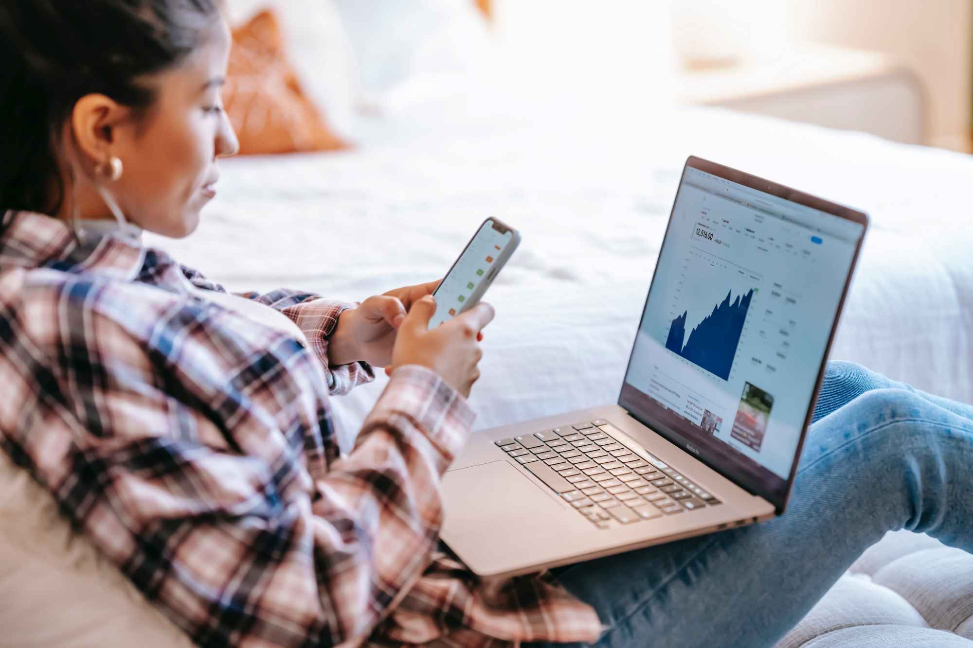 Person in a plaid shirt sitting with laptop on lap displaying a stock market graph and holding a smartphone.