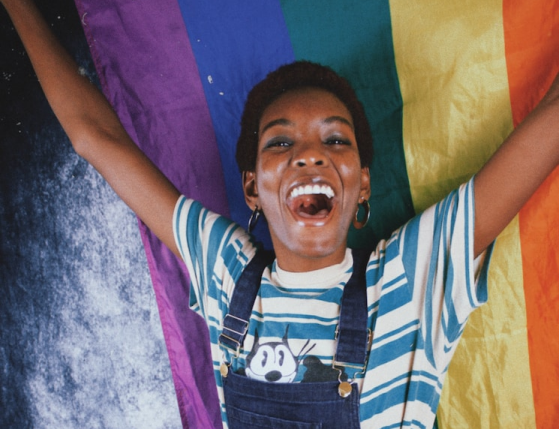 Person joyfully raising their arms in front of a rainbow pride flag.